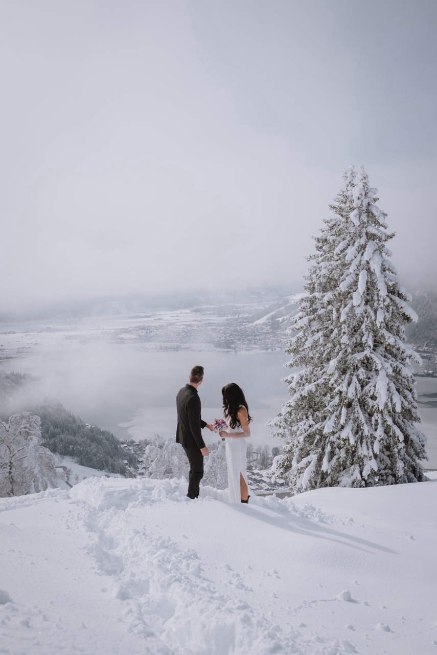 Brautpaar steht Hand in Hand in verschneiter Winterlandschaft mit Blick ins Tal